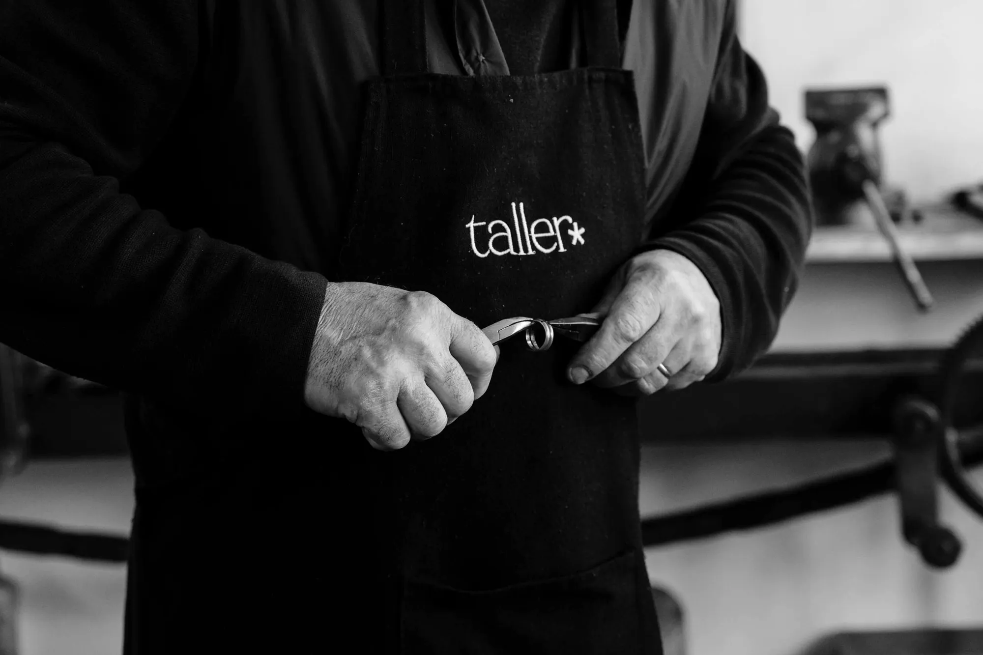 Un joier amb el davantal de taller* sosté una eina petita davant del banc de treball.