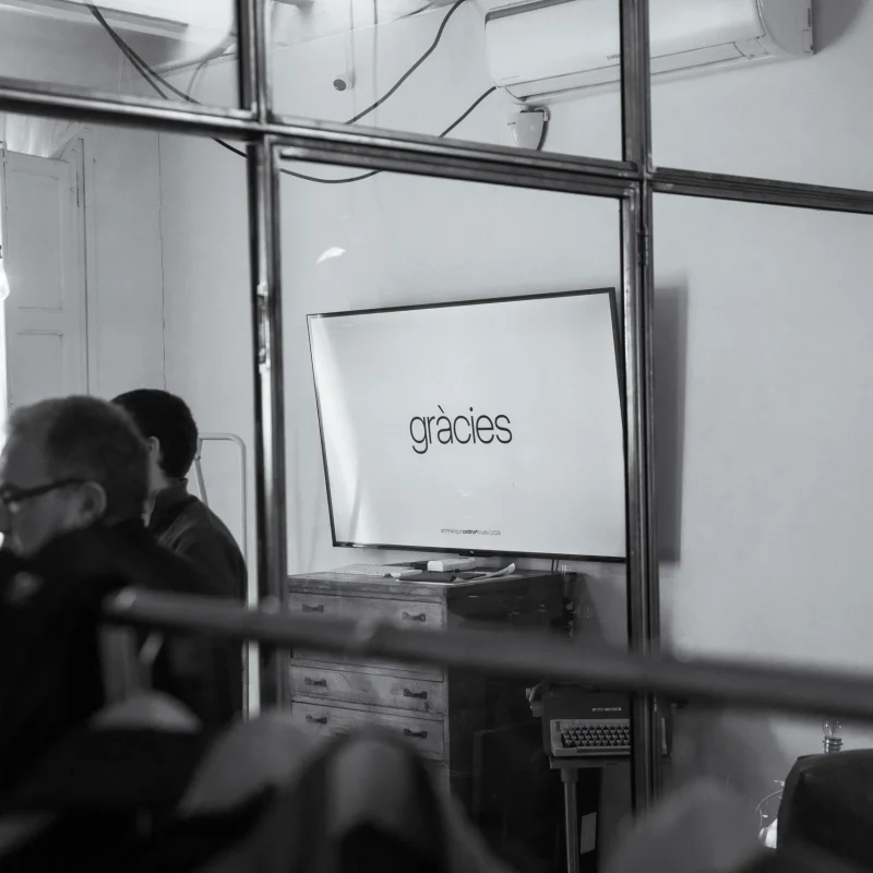 Sala de reuniones del estudio vista desde fuera, a través de una mampara de vidrio y hierro. En la pantalla del fondo, la palabra «gràcies».
