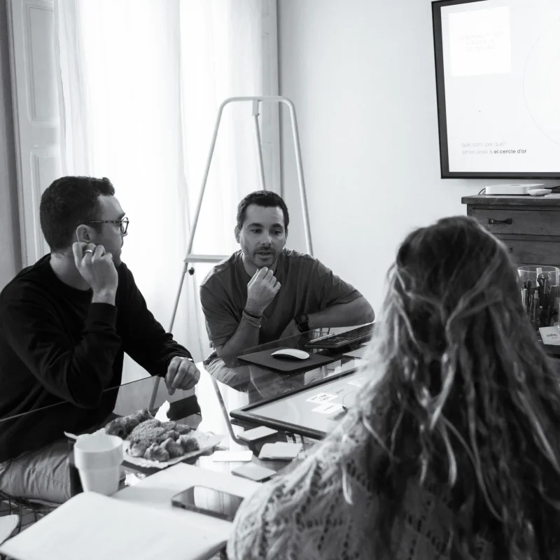 Dos personas conversando alrededor de la mesa de reuniones, con un caballete y una pantalla de presentación al fondo.