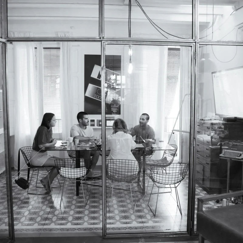 El estudio visto desde fuera de la mampara de vidrio: cuatro personas sentadas alrededor de la mesa redonda, sobre el pavimento hidráulico.